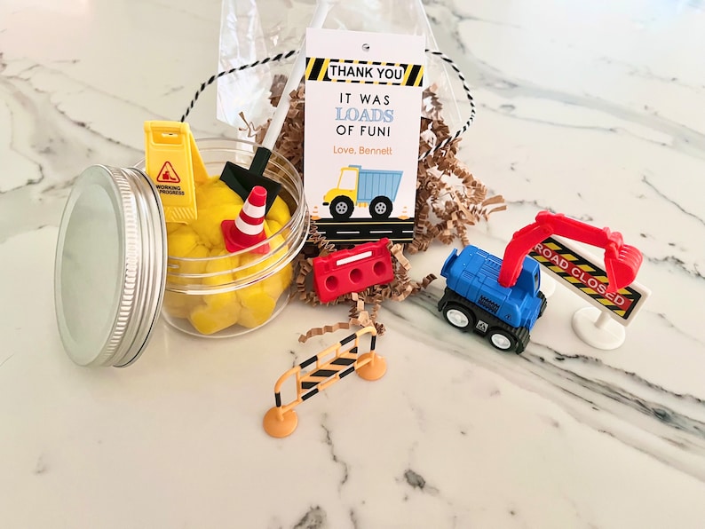 May include: A construction themed party favor with a clear jar filled with yellow play dough, a small toy dump truck, a toy excavator, a toy traffic cone, a toy shovel, a toy road closed sign, and a toy construction barrier. The jar has a silver lid. The favor also includes a thank you tag that reads "Thank You It Was Loads of Fun! Love, Bennett".