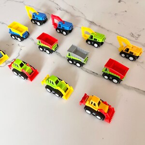 May include: A collection of ten toy construction vehicles, including dump trucks, excavators, and bulldozers. The vehicles are made of plastic and are painted in various colors, including yellow, green, red, blue, and gray.