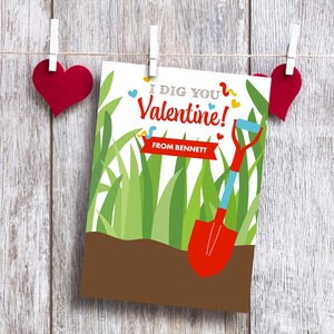 May include: A Valentine's Day card with the message "I DIG YOU Valentine!" and a red shovel illustration. The card is decorated with green grass and a brown soil base. Red heart decorations hang above the card.
