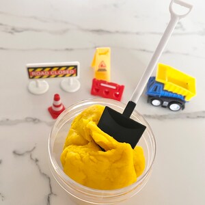 May include: A yellow play dough in a clear container with a black and white toy shovel. The play dough is surrounded by toy construction signs, a toy traffic cone, and a toy dump truck.