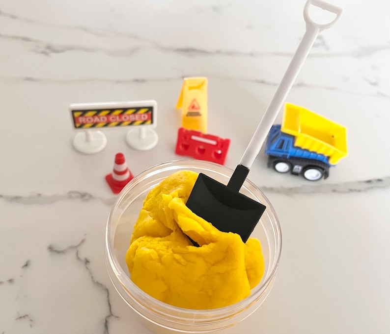 May include: A yellow play dough in a clear container with a black and white toy shovel. The play dough is surrounded by toy construction signs, a toy traffic cone, and a toy dump truck.
