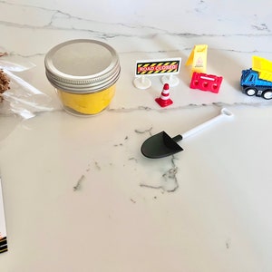 May include: A small jar of yellow play dough with a white lid, a toy construction truck, a toy shovel, a toy traffic cone, a toy road closed sign, and a toy construction barricade. The items are on a white marble surface. The text on the sign says "Road Closed".