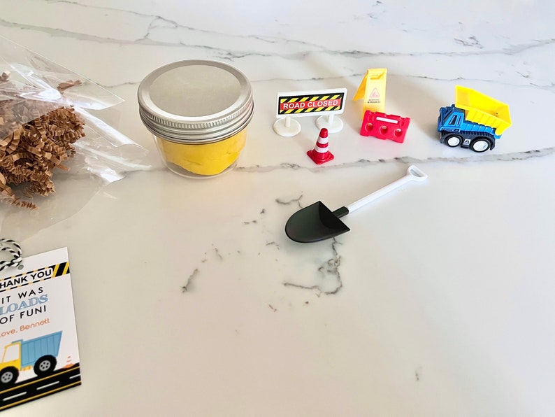 May include: A small jar of yellow play dough with a white lid, a toy construction truck, a toy shovel, a toy traffic cone, a toy road closed sign, and a toy construction barricade. The items are on a white marble surface. The text on the sign says "Road Closed".