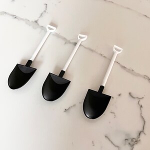 May include: Three black plastic miniature shovels with white handles. The shovels are arranged in a row on a white marble surface.