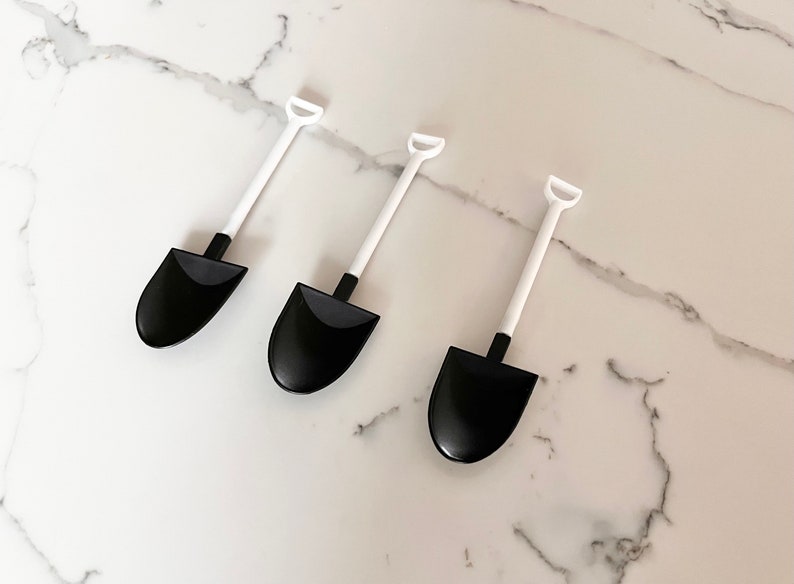 May include: Three black plastic miniature shovels with white handles. The shovels are arranged in a row on a white marble surface.