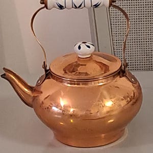 May include: A copper tea kettle with a curved spout and a handle made of copper and white ceramic. The ceramic handle has a blue floral design. The lid also has a white ceramic knob with a blue floral design.