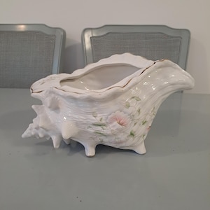 May include: White ceramic seashell planter with a textured surface, featuring pink floral accents and gold trim. The planter has a wide opening and is supported by three small feet. A decorative piece for home decor.