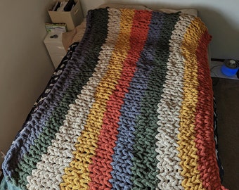 May include: A multi-coloured chunky knit blanket, featuring stripes of grey, green, orange, yellow, and cream. The blanket is displayed on a bed, highlighting its textured appearance. The thick yarn gives a cosy and inviting look.