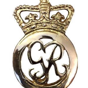 May include: A gold-colored metal badge featuring a crown above a circular emblem. The emblem has the intertwined letters "GR" in a decorative, openwork design. The badge has a polished, reflective surface.