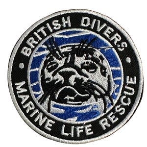 May include: Circular embroidered patch with a seal illustration at the centre. The patch features the text "BRITISH DIVERS MARINE LIFE RESCUE" in white lettering on a black background with blue accents, suitable for application on clothing.