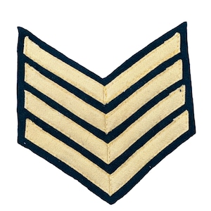May include: A black chevron patch with five gold stripes. The patch is made of fabric and has a stitched edge.