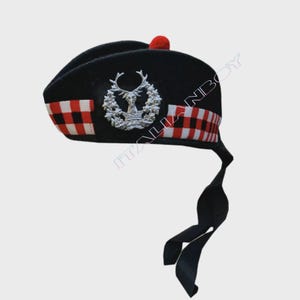 May include: A black Glengarry bonnet, a traditional Scottish hat, with a red pom-pom. It has a silver stag emblem and a red and white checkered band. Black ribbons hang from the back of the hat.