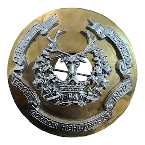 May include: A gold and silver metal badge with the words "Gordon Highlanders" around the edge. The centre features a stag's head with a crown and the words "Peninsula, Waterloo, Egypt, India".