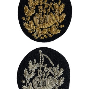 Puede incluir: Dos parches redondos de fieltro negro con gaitas bordadas en oro y plata. Las gaitas están rodeadas por una corona de hojas.