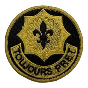 Könnte beinhalten: Ein runder, gestickter Aufnäher in Schwarz und Gold mit einer Fleur-de-lis in der Mitte und dem Text "TOUJOURS PRET" am Rand.