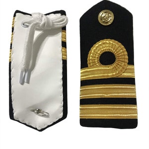 Könnte beinhalten: Ein Paar goldene und schwarze Schulterklappen mit vier goldenen Streifen und einem goldenen runden Emblem. Die Schulterklappen sind an einer weißen Unterlage mit einem weißen Kordel befestigt.