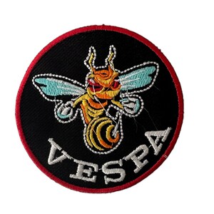 Può includere: Una toppa circolare ricamata di colore nero e rosso con un'ape gialla e arancione con ali blu e la parola "VESPA" in bianco.