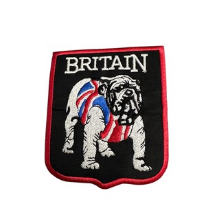 Puede incluir: Parche bordado con un diseño de bulldog y la bandera de la Union Jack. El parche tiene un fondo negro con la palabra "BRITAIN" en blanco. El borde es rojo.