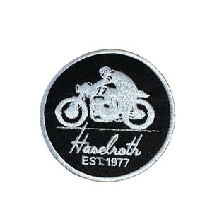 Może przedstawiać: Okrągła, czarno-biała naszywka z haftem przedstawiającym motocykl i motocyklistę. Projekt zawiera napis "Haselroth" w ozdobnej czcionce i "EST.1977" mniejszym fontem, z numerem "77" na motocyklu.