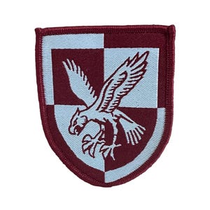 Peut inclure: Écusson brodé bordeaux et blanc représentant un aigle aux ailes déployées. L'écusson est en forme de bouclier, avec un motif à carreaux en arrière-plan. L'aigle est blanc et le bord est bordeaux.