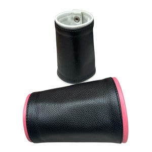 May include: Two black leather wrist cuffs with white and pink accents. One cuff is upright, showing a white interior and a snap closure. The other cuff is lying down, displaying a pink edge.