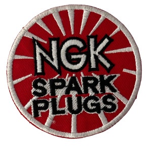 May include: A red and white embroidered patch with the text "NGK SPARK PLUGS" in black lettering.