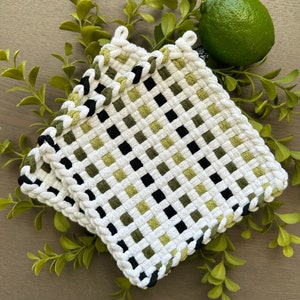 Puede incluir: Dos agarraderas blancas tejidas con un patrón de tablero de ajedrez de cuadrados negros y verdes. Los bordes están adornados con un borde trenzado. Una lima verde se encuentra junto a las agarraderas, con ramitas verdes en el fondo.