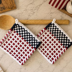 May include: Two square potholders featuring a woven grid pattern in red, white, and navy blue yarn. Each potholder has a white crocheted border and a small loop for hanging. The design mimics a flag pattern with a solid blue corner section.