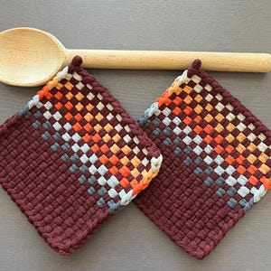 May include: Two burgundy woven potholders with a diagonal pattern of orange, grey, and white squares. A wooden spoon rests above the potholders. These kitchen accessories are ideal for protecting surfaces from heat.