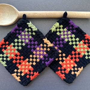 Puede incluir: Dos salvamanteles cuadrados negros tejidos con patrones geométricos naranjas, morados y verdes. Una cuchara de madera descansa sobre los salvamanteles. Los salvamanteles tienen un lazo para colgar.