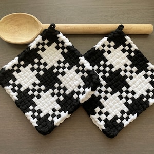 May include: Two black and white woven pot holders, made with thick yarn, are laying on a gray surface. A wooden spoon is laying on top of the pot holders.