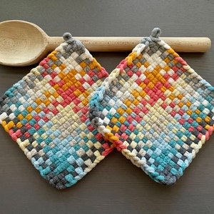 May include: Two square, woven potholders in a colorful pattern of gray, blue, yellow, orange, and pink. A wooden spoon rests behind the potholders. The potholders have a loop for hanging.