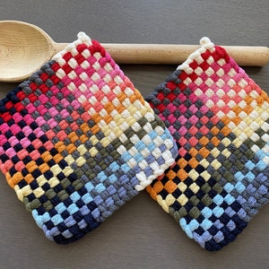 May include: Two colorful woven potholders in a diamond pattern, featuring a gradient of colors from red to blue. A wooden spoon rests above the potholders, adding a rustic touch. The potholders are approximately 7 inches square.