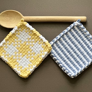 May include: Two woven potholders, one yellow and white checkered, the other blue and white striped, with white edging. A wooden spoon rests above them on a brown surface. Kitchen textiles.