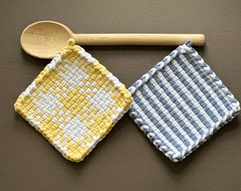 Pode incluir: Duas pegas tecidas, uma xadrez amarela e branca, a outra listrada azul e branca, com bordas brancas. Uma colher de pau repousa sobre elas em uma superfície marrom. Têxteis de cozinha.
