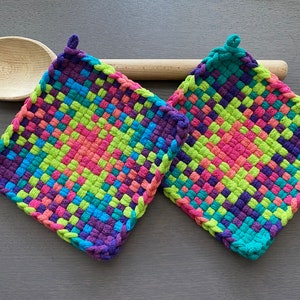 Può includere: Due presine quadrate, tessute in colori vivaci: rosa, verde, blu e viola. Ogni presina ha un anello per appenderla. Un cucchiaio di legno e un mattarello sono sullo sfondo.