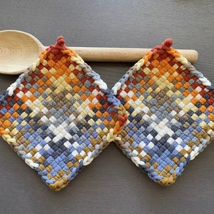 Op de afbeelding: Twee geweven pannenlappen in een ruitpatroon met een kleurengradient van oranje, blauw, wit en bruin. Een houten lepel ligt boven de pannenlappen. Deze keukenaccessoires zijn ontworpen voor hittebestendigheid.