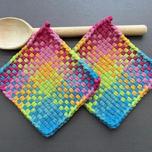 May include: Two colorful woven potholders in shades of pink, blue, green, and yellow. A wooden spoon rests behind the potholders on a dark gray surface. The potholders are square-shaped with a braided edge.