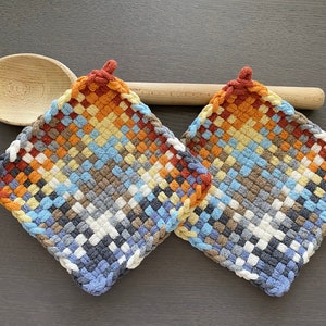 May include: Two square woven potholders in a colourful pattern of blue, orange, brown, and white. Each potholder has a small loop at the top. A wooden spoon rests behind the potholders.