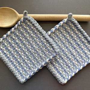 May include: Two square, woven potholders in shades of blue, gray, and white, with a braided gray border. A wooden spoon rests above the potholders. The potholders are designed for kitchen use.
