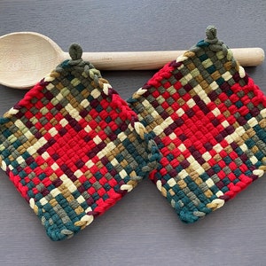 May include: Two woven potholders with a red and plaid design, featuring green, beige, and burgundy hues. A wooden spoon rests behind the potholders. The potholders have a loop for hanging.