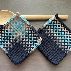 Puede incluir: Dos salvamanteles cuadrados tejidos en tonos azules, grises y beige. Los salvamanteles tienen un borde trenzado y un lazo para colgar. Una cuchara de madera descansa detrás de ellos sobre una superficie gris.