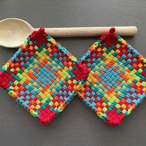 Puede incluir: Dos salvamanteles tejidos coloridos con un borde rojo y un patrón tejido de azul, verde, naranja y amarillo. Una cuchara de madera descansa sobre los salvamanteles sobre una superficie gris. Los salvamanteles son cuadrados.