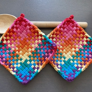 May include: Two diamond-shaped woven potholders in a vibrant gradient of colours, including red, purple, orange, yellow, and blue. Each potholder has a small loop at the top. A wooden spoon is positioned behind them.
