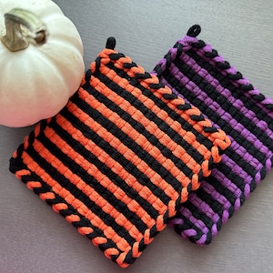 Könnte beinhalten: Zwei quadratische Topflappen mit schwarz-orangen Streifen und schwarz-violetten Streifen. Die Topflappen haben eine geflochtene schwarze Bordüre und eine Schlaufe zum Aufhängen. Im Hintergrund ist ein weißer Kürbis.