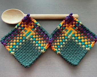 May include: Two square woven potholders with a gradient of colors, including green, orange, yellow, purple, and teal. A wooden spoon rests behind the potholders. The potholders are made of a thick, textured material.