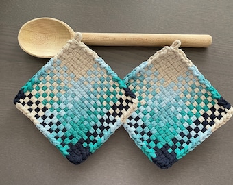Pode incluir: Duas pegas quadradas, tecidas com um gradiente de cores do azul marinho ao azul claro e bege. Uma colher de pau repousa sobre as pegas numa superfície cinzenta. As pegas têm uma argola para pendurar.