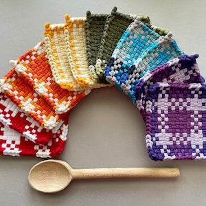 Puede incluir: Un conjunto de agarraderas de olla de crochet de colores en un patrón de arcoíris, dispuestas en semicírculo con una cuchara de madera en la parte delantera.