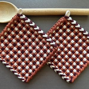 Könnte beinhalten: Zwei quadratische, gewebte Topflappen in einem Schachbrettmuster aus Rost, Weiß und Burgunderrot. Die Kanten sind mit einem geflochtenen Rand versehen. Ein Holzlöffel liegt über den Topflappen.
