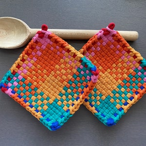 May include: Two woven potholders in vibrant colours of orange, pink, teal, and blue. Each square-shaped holder has a loop for hanging and a woven design. A wooden spoon rests behind the potholders.